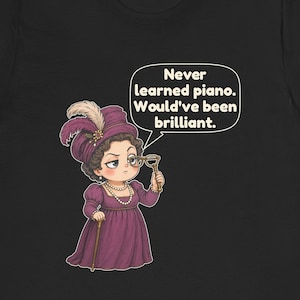 May include: A black t-shirt featuring a cartoon illustration of a person in a purple Victorian-style dress and hat, holding a monocle. A speech bubble above contains the text: Never learned piano. Would've been brilliant.