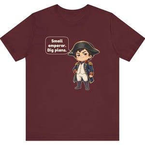 May include: A maroon t-shirt featuring a cartoon illustration of a historical figure in a blue military uniform and bicorne hat, with a speech bubble containing the text: Small emperor. Big plans.