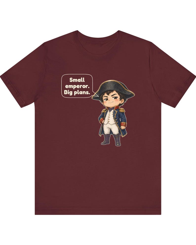 May include: A maroon t-shirt featuring a cartoon illustration of a historical figure in a blue military uniform and bicorne hat, with a speech bubble containing the text: Small emperor. Big plans.