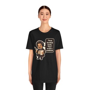 May include: A black t-shirt featuring a cartoon illustration of a person with an afro, wearing a suit and holding a quill and book. A speech bubble contains the text: Three hundred books. Then I wrote a cookbook.