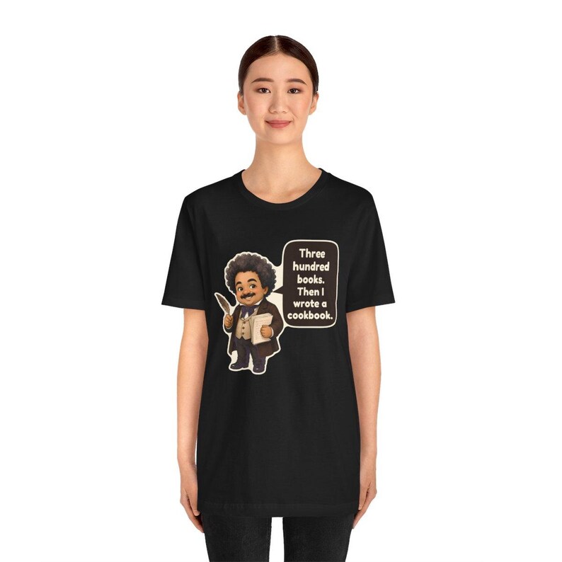 May include: A black t-shirt featuring a cartoon illustration of a person with an afro, wearing a suit and holding a quill and book. A speech bubble contains the text: Three hundred books. Then I wrote a cookbook.