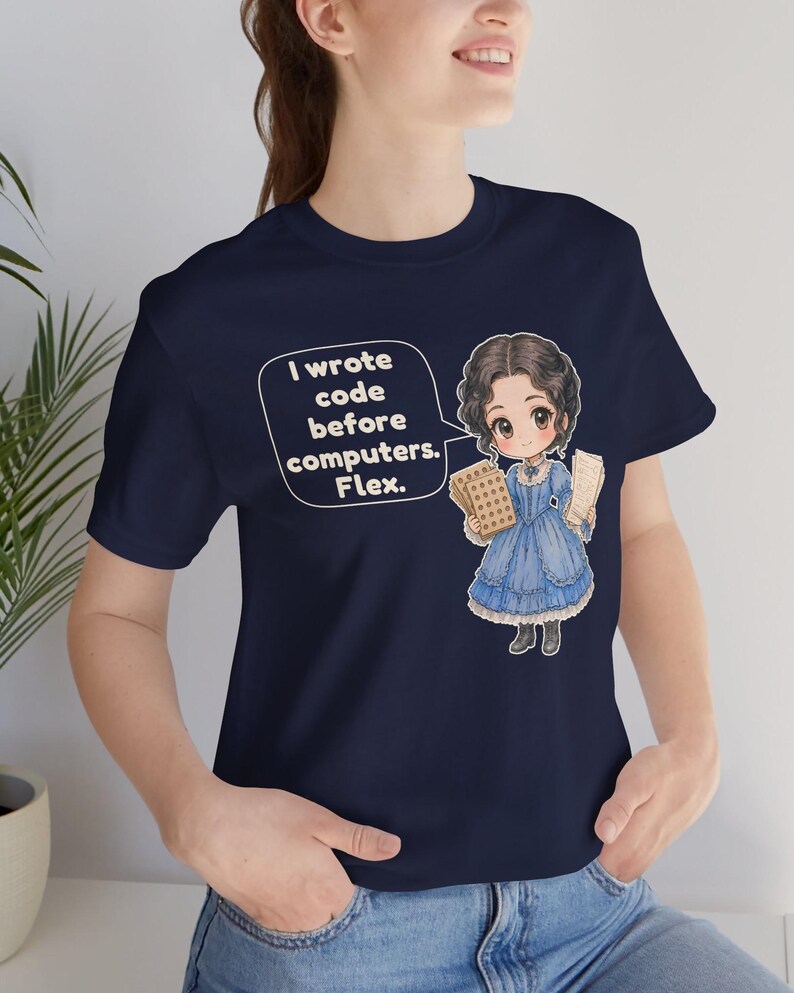 May include: A navy blue t-shirt featuring a graphic of a person in a blue Victorian-style dress holding punch cards. A speech bubble next to the character contains the text: I wrote code before computers. Flex.