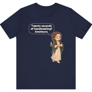 Op de afbeelding: Een marineblauw t-shirt met een cartoonillustratie van een persoon met bruin haar in een vlecht, gekleed in een cr&egrave;mekleurige jurk en donkere sjaal, met een brandende kaars. Een tekstballon bevat de tekst: Twenty seconds of handwashing? Amateurs.