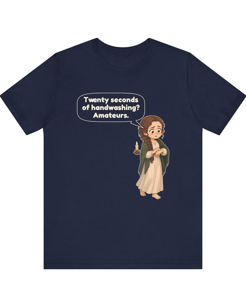 Op de afbeelding: Een marineblauw t-shirt met een cartoonillustratie van een persoon met bruin haar in een vlecht, gekleed in een cr&egrave;mekleurige jurk en donkere sjaal, met een brandende kaars. Een tekstballon bevat de tekst: Twenty seconds of handwashing? Amateurs.