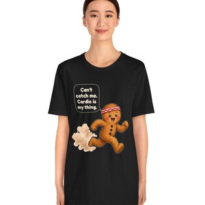 May include: A black t-shirt featuring a graphic of a running gingerbread man wearing a red and white headband. The gingerbread man has a speech bubble that says, "Can't catch me. Cardio is my thing." A cloud of dust trails behind the figure.