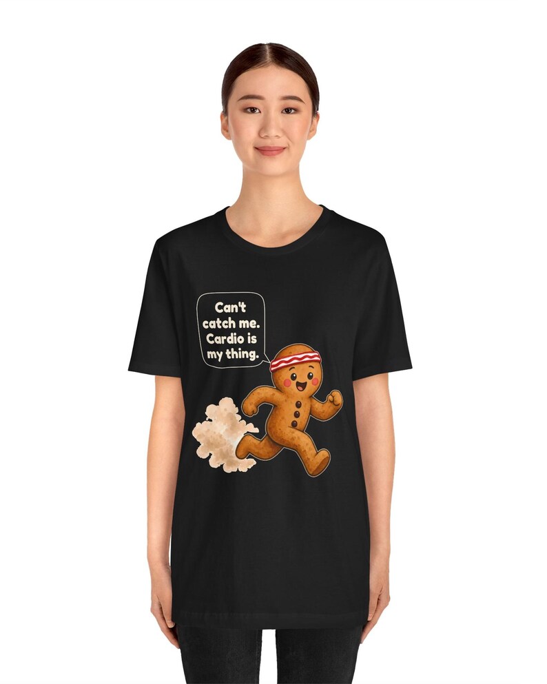 May include: A black t-shirt featuring a graphic of a running gingerbread man wearing a red and white headband. The gingerbread man has a speech bubble that says, "Can't catch me. Cardio is my thing." A cloud of dust trails behind the figure.