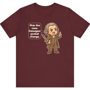 May include: A maroon short-sleeve t-shirt featuring a cartoon illustration of a lawyer in 18th-century attire holding a quill. A speech bubble contains the text: Won the case. Damages: pocket change.