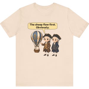 May include: A light beige t-shirt featuring a graphic of two figures in 18th-century attire standing next to a hot air balloon with a sheep in the basket. A speech bubble above them contains the text: The sheep flew first. Obviously.