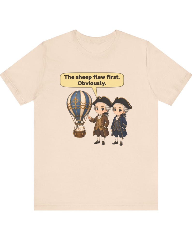 May include: A light beige t-shirt featuring a graphic of two figures in 18th-century attire standing next to a hot air balloon with a sheep in the basket. A speech bubble above them contains the text: The sheep flew first. Obviously.