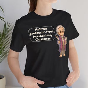 Pu&ograve; includere: Una t-shirt nera a maniche corte con un'illustrazione a cartoni animati di un uomo con capelli bianchi e occhiali che tiene in mano una penna e una pergamena. Un fumetto contiene il testo: Hebrew professor. Poet. Accidentally Christmas.
