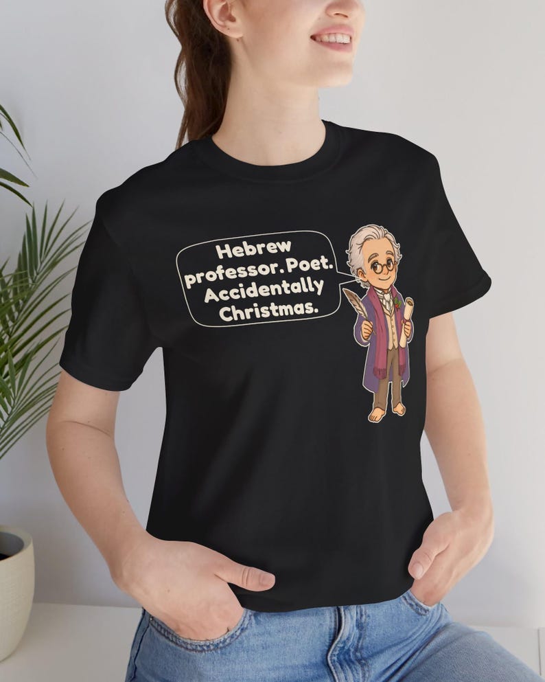 Pu&ograve; includere: Una t-shirt nera a maniche corte con un'illustrazione a cartoni animati di un uomo con capelli bianchi e occhiali che tiene in mano una penna e una pergamena. Un fumetto contiene il testo: Hebrew professor. Poet. Accidentally Christmas.