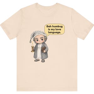 Puede incluir: Una camiseta de color beige claro con una ilustraci&oacute;n de dibujos animados de un personaje gru&ntilde;&oacute;n con gorro de dormir y bata, sosteniendo una vela y un libro. Un bocadillo junto al personaje contiene el texto: Bah humbug is my love language.