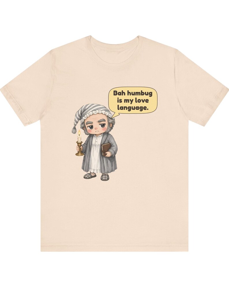 Puede incluir: Una camiseta de color beige claro con una ilustraci&oacute;n de dibujos animados de un personaje gru&ntilde;&oacute;n con gorro de dormir y bata, sosteniendo una vela y un libro. Un bocadillo junto al personaje contiene el texto: Bah humbug is my love language.
