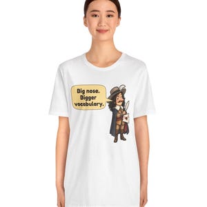 May include: A white short-sleeve t-shirt featuring a cartoon illustration of a historical figure with a large nose, wearing a hat and cape, holding a quill and letter. A yellow speech bubble contains the text: Big nose. Bigger vocabulary.