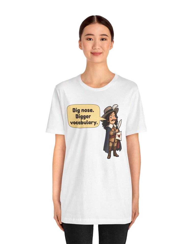 May include: A white short-sleeve t-shirt featuring a cartoon illustration of a historical figure with a large nose, wearing a hat and cape, holding a quill and letter. A yellow speech bubble contains the text: Big nose. Bigger vocabulary.