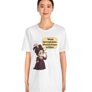 May include: A white short-sleeve t-shirt featuring a graphic of a person in a purple Victorian-style dress and hat, holding a monocle. A speech bubble contains the text: Never learned piano. Would've been brilliant.