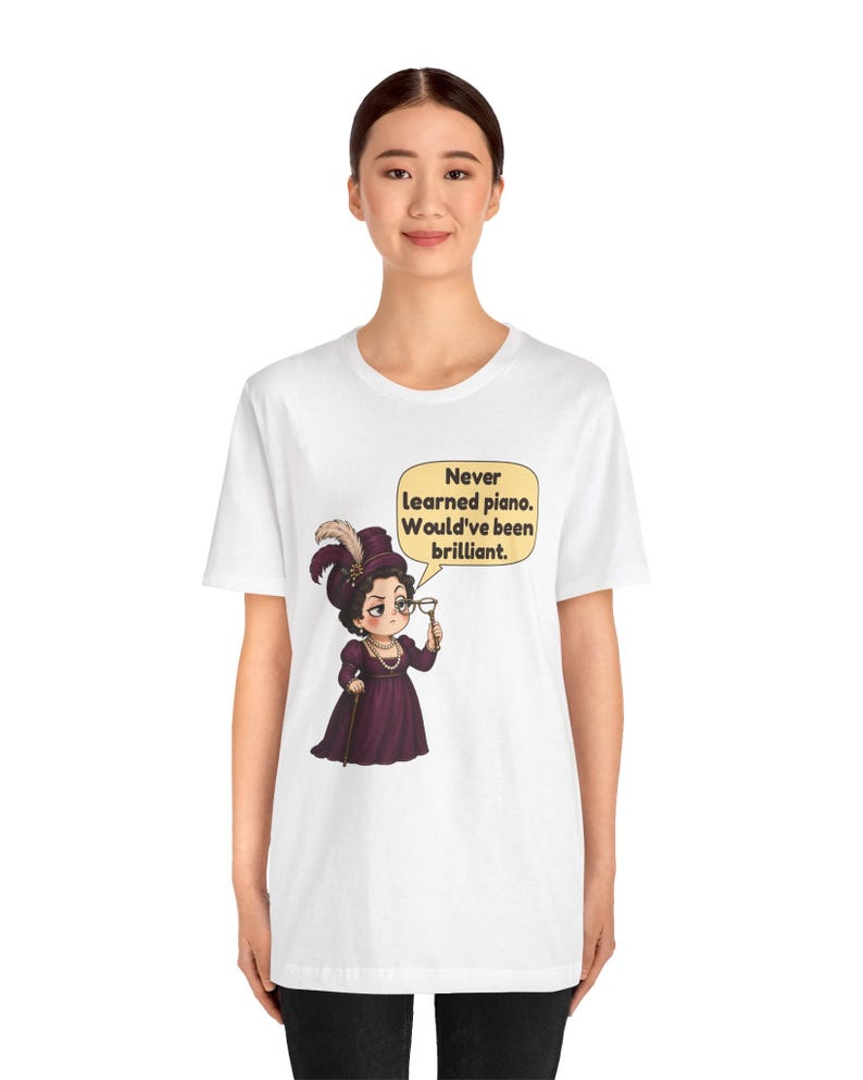 May include: A white short-sleeve t-shirt featuring a graphic of a person in a purple Victorian-style dress and hat, holding a monocle. A speech bubble contains the text: Never learned piano. Would've been brilliant.