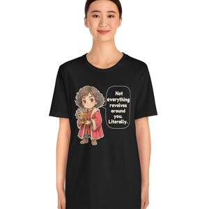 May include: A black short-sleeve t-shirt featuring a cartoon illustration of a person with curly brown hair wearing a red coat and holding a golden armillary sphere. A speech bubble contains the text: Not everything revolves around you. Literally.