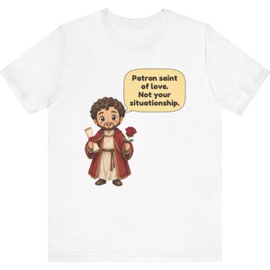 Puede incluir: Una camiseta blanca con una ilustraci&oacute;n de dibujos animados de una figura barbuda con t&uacute;nica roja que sostiene un pergamino y una rosa roja. Un globo de texto junto a la figura contiene el texto: Patron saint of love. Not your situationship.