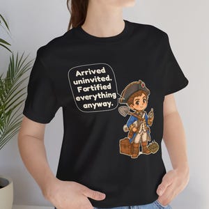 May include: A black t-shirt featuring a cartoon illustration of a historical figure in a blue coat and tricorn hat holding a shovel and compass, with a speech bubble containing the text: Arrived uninvited. Fortified everything anyway.