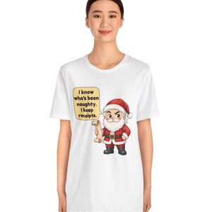 May include: A white t-shirt featuring a cartoon illustration of Santa Claus wearing a red suit and hat, holding a scroll. A yellow speech bubble next to him contains the black text: I know who's been naughty. I keep receipts.