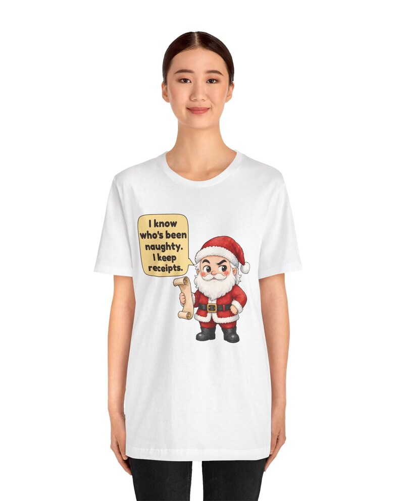 May include: A white t-shirt featuring a cartoon illustration of Santa Claus wearing a red suit and hat, holding a scroll. A yellow speech bubble next to him contains the black text: I know who's been naughty. I keep receipts.
