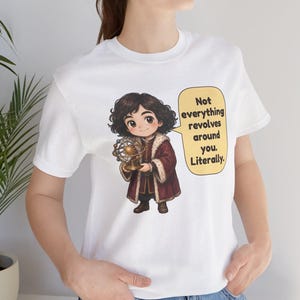 May include: A white t-shirt featuring a cartoon illustration of a person with dark curly hair wearing a maroon coat with fur trim, holding a gold armillary sphere. A yellow speech bubble contains the text: Not everything revolves around you. Literally.