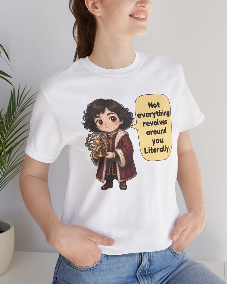 May include: A white t-shirt featuring a cartoon illustration of a person with dark curly hair wearing a maroon coat with fur trim, holding a gold armillary sphere. A yellow speech bubble contains the text: Not everything revolves around you. Literally.