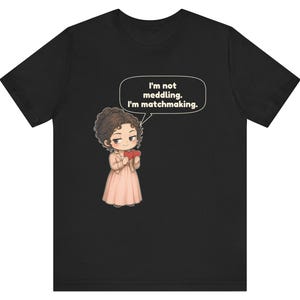 K&ouml;nnte beinhalten: Ein schwarzes T-Shirt mit einer Cartoon-Illustration einer Person mit braunen Haaren, die ein hellrosa Kleid und eine Jacke tr&auml;gt und zwei rote Herzen h&auml;lt. Eine Sprechblase enth&auml;lt den Text, I'm not meddling. I'm matchmaking.
