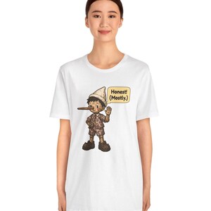 K&ouml;nnte beinhalten: Ein wei&szlig;es Kurzarm-T-Shirt mit einer Grafik einer Holzpuppe mit langer Nase, die einen Hut und eine kurze Hose tr&auml;gt. Die Figur winkt mit einer Hand und eine Sprechblase neben ihr enth&auml;lt den Text: Honest! (Mostly.)