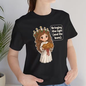 May include: A black short-sleeve t-shirt featuring a cartoon illustration of a person with long brown hair wearing a white robe and a candle crown. The person holds a tray of buns, with a speech bubble that reads: Bringing the light (and the buns).