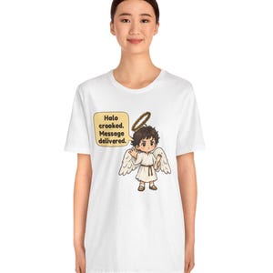 May include: A white t-shirt featuring a graphic of a cartoon angel with brown hair, white wings, and a halo. The angel holds a scroll and a speech bubble reads: Halo crooked. Message delivered. The design is centered on the chest area.