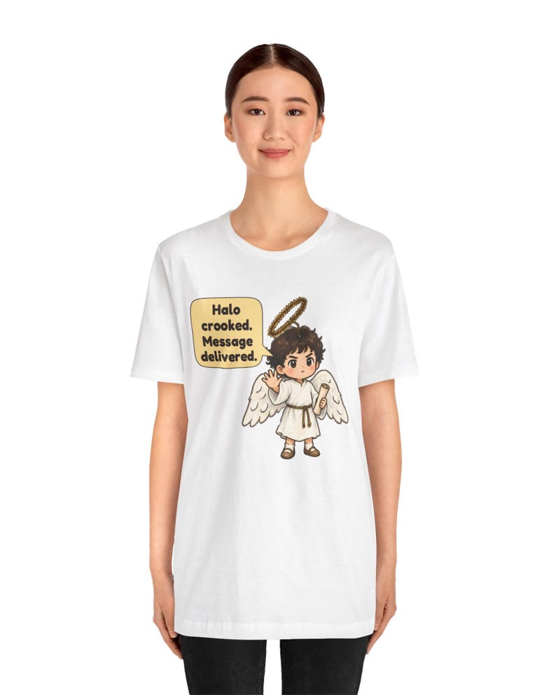 May include: A white t-shirt featuring a graphic of a cartoon angel with brown hair, white wings, and a halo. The angel holds a scroll and a speech bubble reads: Halo crooked. Message delivered. The design is centered on the chest area.