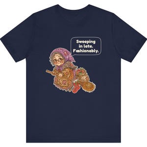 Puede incluir: Una camiseta azul marino de manga corta con un gr&aacute;fico de una persona mayor con pa&ntilde;uelo y gafas montada en una escoba. El gr&aacute;fico incluye un bocadillo con el texto "Sweeping in late. Fashionably." y un saco con varios objetos.