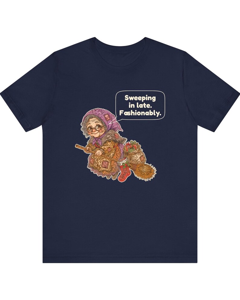 Puede incluir: Una camiseta azul marino de manga corta con un gr&aacute;fico de una persona mayor con pa&ntilde;uelo y gafas montada en una escoba. El gr&aacute;fico incluye un bocadillo con el texto "Sweeping in late. Fashionably." y un saco con varios objetos.