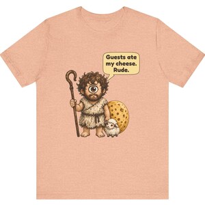 May include: A peach-colored t-shirt featuring a cartoon cyclops holding a wooden staff and standing next to a small sheep and a large piece of Swiss cheese. A speech bubble above the cyclops contains the text: Guests ate my cheese. Rude.