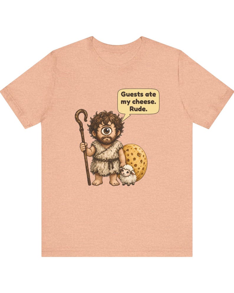 May include: A peach-colored t-shirt featuring a cartoon cyclops holding a wooden staff and standing next to a small sheep and a large piece of Swiss cheese. A speech bubble above the cyclops contains the text: Guests ate my cheese. Rude.