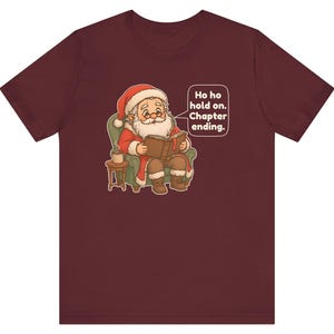 Op de afbeelding: Een bordeauxrood t-shirt met korte mouwen en een afbeelding van de Kerstman die in een groene fauteuil zit en een boek leest. Een tekstballon ernaast bevat de tekst: Ho ho hold on. Chapter ending. Een klein bijzettafeltje met mok staat ernaast.