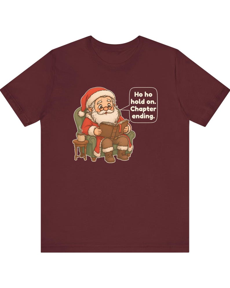 Op de afbeelding: Een bordeauxrood t-shirt met korte mouwen en een afbeelding van de Kerstman die in een groene fauteuil zit en een boek leest. Een tekstballon ernaast bevat de tekst: Ho ho hold on. Chapter ending. Een klein bijzettafeltje met mok staat ernaast.