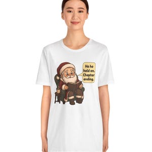 Op de afbeelding: Een wit t-shirt met een afbeelding van de Kerstman die in een groene fauteuil zit en een boek leest, met een mok op een bijzettafel. Een tekstballon naast hem bevat de tekst: Ho ho hold on. Chapter ending.