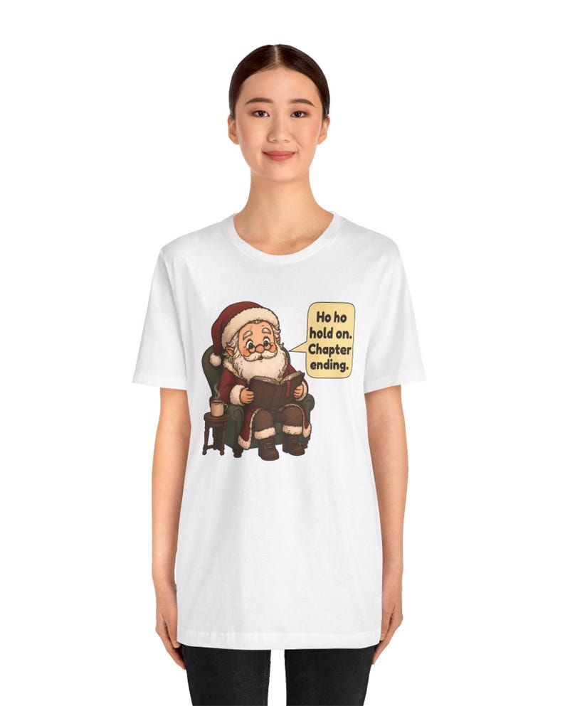 Op de afbeelding: Een wit t-shirt met een afbeelding van de Kerstman die in een groene fauteuil zit en een boek leest, met een mok op een bijzettafel. Een tekstballon naast hem bevat de tekst: Ho ho hold on. Chapter ending.