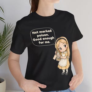 May include: A black t-shirt featuring a cartoon illustration of a blonde girl in a yellow dress and white apron holding a glass bottle. A speech bubble above her contains the text: Not marked poison. Good enough for me.