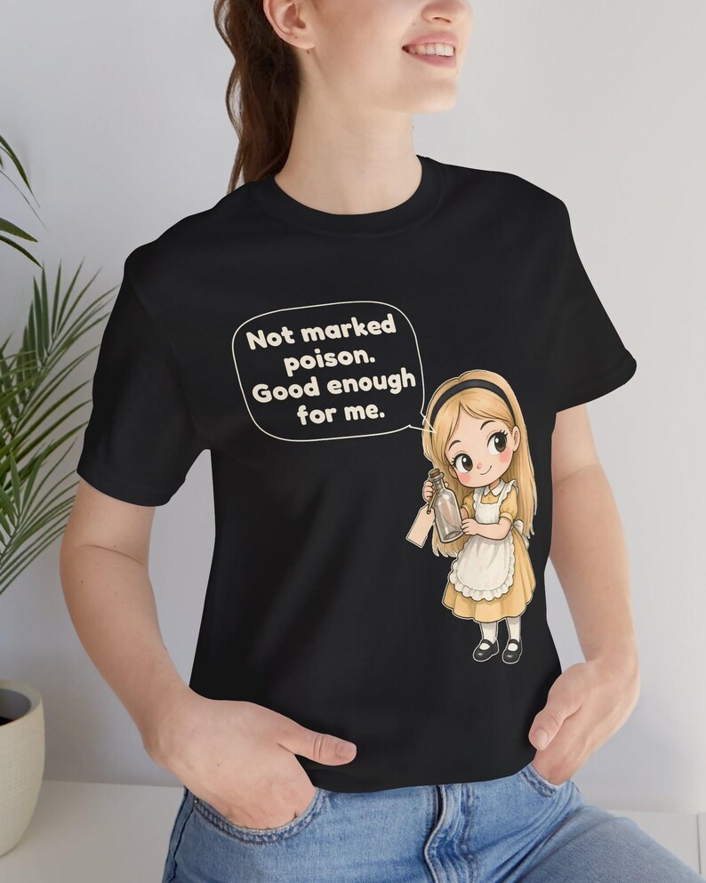 May include: A black t-shirt featuring a cartoon illustration of a blonde girl in a yellow dress and white apron holding a glass bottle. A speech bubble above her contains the text: Not marked poison. Good enough for me.