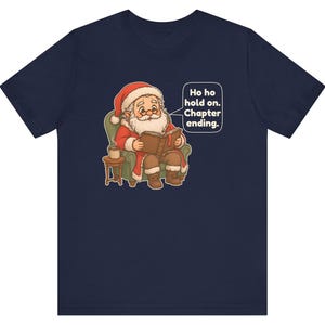 Op de afbeelding: Een marineblauw t-shirt met een afbeelding van de Kerstman die in een groene fauteuil zit, een boek leest en een mok op een bijzettafel heeft staan. Een tekstballon bevat de tekst: Ho ho hold on. Chapter ending.