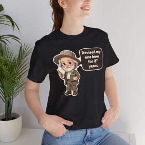 May include: A black t-shirt featuring a cartoon illustration of an elderly man with a white beard wearing a brown hat and suit. He holds a small book and a pen. A speech bubble next to him contains the text: Revised my one book for 37 years.