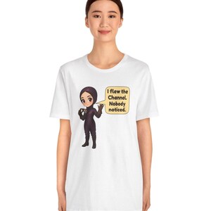 Puede incluir: Una camiseta blanca de manga corta con una ilustraci&oacute;n de dibujos animados de un personaje con traje de vuelo y casco de color morado oscuro. El personaje sostiene unas gafas y un bocadillo que dice, I flew the Channel. Nobody noticed.