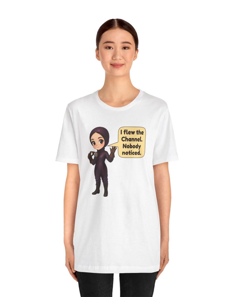 Puede incluir: Una camiseta blanca de manga corta con una ilustraci&oacute;n de dibujos animados de un personaje con traje de vuelo y casco de color morado oscuro. El personaje sostiene unas gafas y un bocadillo que dice, I flew the Channel. Nobody noticed.