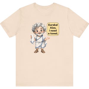 May include: A light beige t-shirt featuring a cartoon illustration of a bearded man in a white toga and brown sandals holding a white towel. A yellow speech bubble next to the character contains the black text: Eureka! Also, I need a towel.