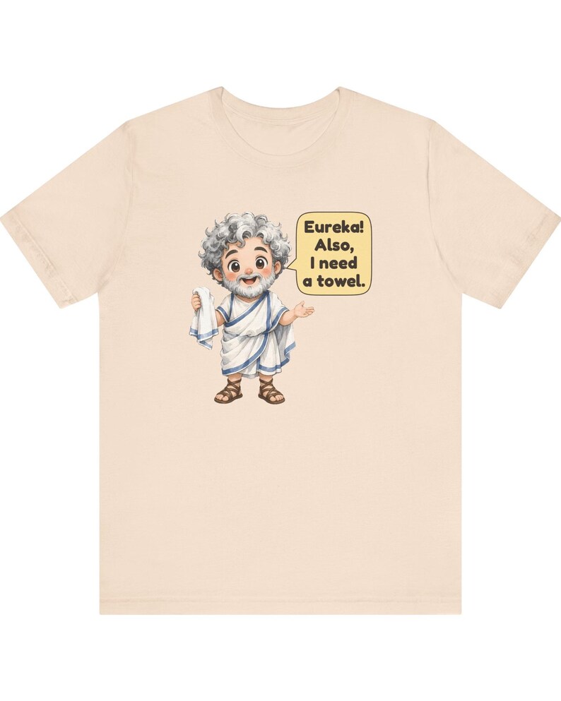 May include: A light beige t-shirt featuring a cartoon illustration of a bearded man in a white toga and brown sandals holding a white towel. A yellow speech bubble next to the character contains the black text: Eureka! Also, I need a towel.