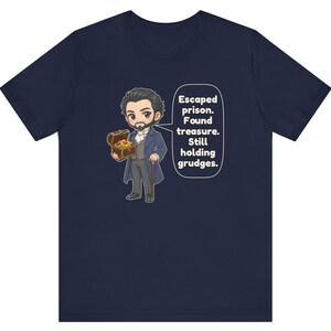 May include: A navy blue t-shirt featuring a cartoon illustration of a bearded man in a formal coat holding a treasure chest. A speech bubble next to him contains the text: Escaped prison. Found treasure. Still holding grudges.